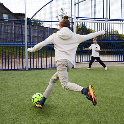 A child is about to score a goal, he is about to hit the football, the goalie in the background is ready. They are playing at a public sport court.